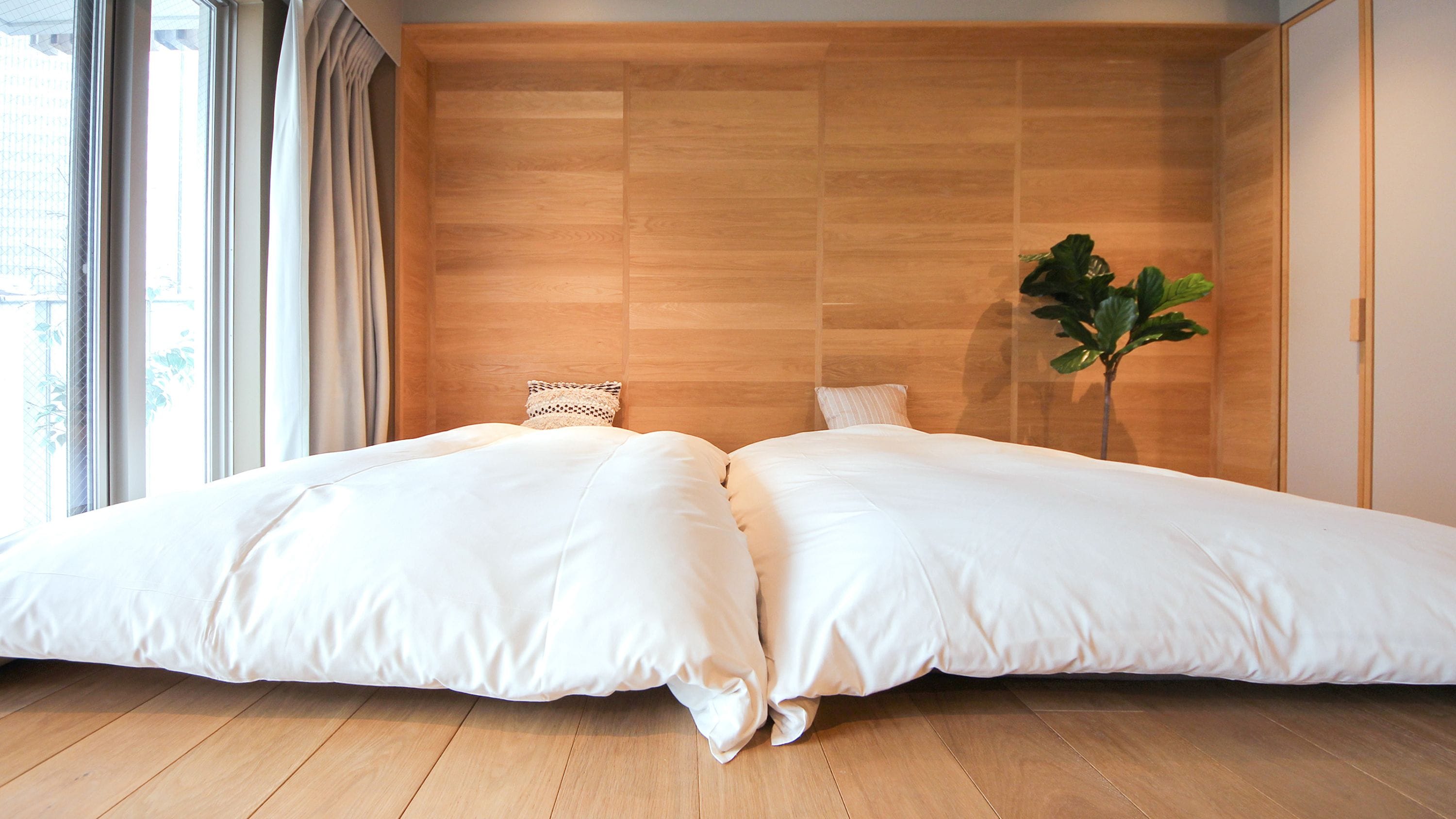 A double bed with white linens in a room with wooden flooring and a large window.