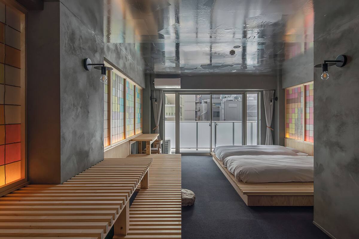 A modern hotel room with a minimalist design, featuring a large bed, a small desk, and a view through a glass door.