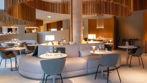 A modern restaurant with round tables and grey upholstered seating, featuring a central wooden column and a ceiling with wooden slats.