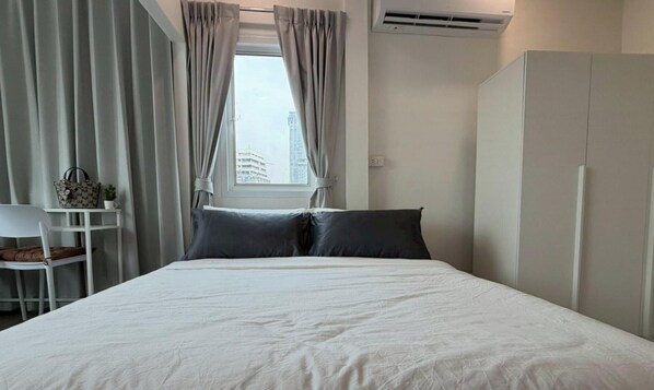 A neatly made bed with white bedding, a white wardrobe, and a window with curtains. - NamasStay Silom (Bangkok)