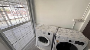 A laundry area with two front-loading washing machines, a window with a metal grid, and a white wall with electrical outlets. - NamasStay Silom (Bangkok)