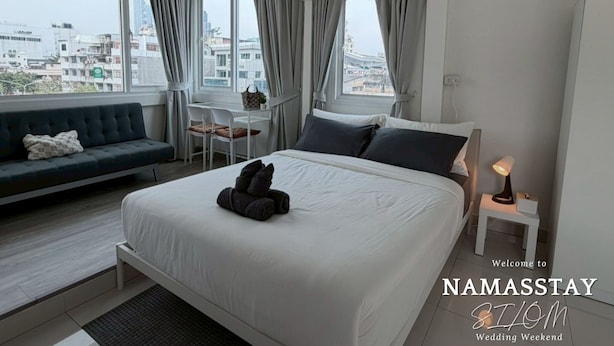 A modern hotel room with a large bed, a sofa, a small table, and a view of the city. - NamasStay Silom (Bangkok)