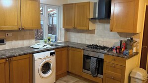 A compact kitchen with wooden cabinets, a washing machine, a stove, and a trash bin. - 2 bed-Sleeps 4-Private Patio-Pets-Near Heathrow (Hounslow)