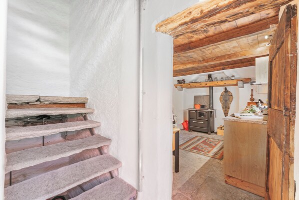 A stone staircase leading to a wooden door, a kitchen area with a rug, and a shelf with jars.