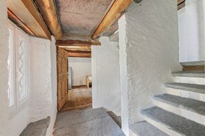 Ein schmaler Korridor mit weißen Wänden und Holzbalken, der zu einer Holztür und einer Treppe mit Steinstufen führt.