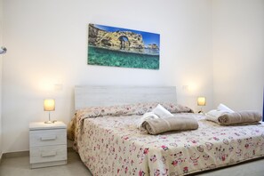 A bedroom with a bed, two bedside lamps, a nightstand, and a wall-mounted painting of a coastal scene.
