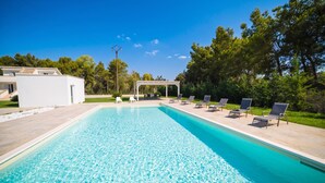 Una piscina rectangular con agua azul cristalina, rodeada por una terraza blanca. Hay sillas de descanso alineadas a lo largo del borde de la piscina, y una zona de estar cubierta con mesas y sillas al fondo.