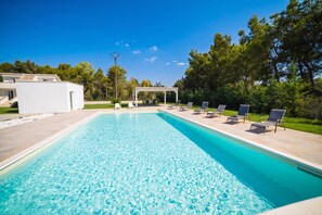 A rectangular swimming pool with clear blue water, surrounded by a white deck. There are lounge chairs aligned along the pool's edge, and a covered seating area with tables and chairs in the background. - Dimora Cristal With Pool And Padel, Galatina, Italy (Galatina)