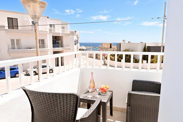A balcony with wicker furniture, a table with a bottle and glasses, and a view of the sea and buildings.