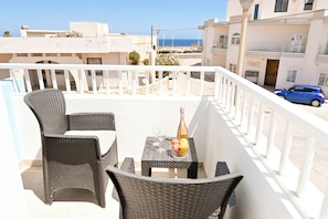 Een balkon met rieten meubilair, een tafel met een fles en fruit, en uitzicht op zee en gebouwen.