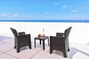 Two wicker chairs and a small table with a bottle and fruit on a terrace with a view of the sea.