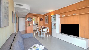 A modern living room with a sofa, a dining table with chairs, a flat-screen TV mounted on a wooden cabinet, and a kitchen area in the background. - Apartment Sea View Panorama, Roquebrune-Cap-Martin, France (Roquebrune-Cap-Martin)