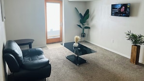 A living room with a black leather sofa, a glass coffee table, a potted plant, and a TV mounted on the wall. - Cozy Hideaway Parks + Trails Near SLC Airport (Lake Point)
