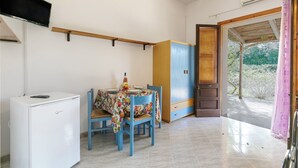 Una cuina compacta amb zona de menjador, un refrigerador blanc, una taula coberta amb un mantell floral i vista a l'exterior per una porta oberta.