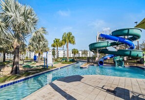 A swimming pool with a water slide, palm trees, and a building in the background.
