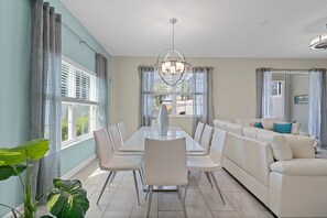 A dining area with a white table and chairs, a chandelier, and a view of the outdoors through large windows.