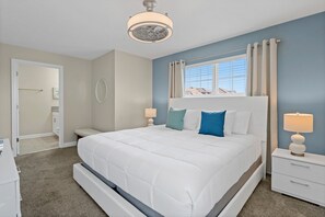 A bedroom with a large bed, bedside tables, a ceiling fan, a window with curtains, and a door leading to another room.