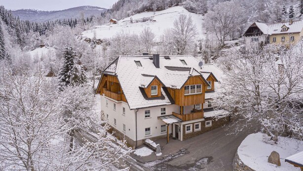 Una casa grande de madera, de varios pisos y con techo inclinado, rodeada de árboles cubiertos de nieve y un paisaje montañoso al fondo.