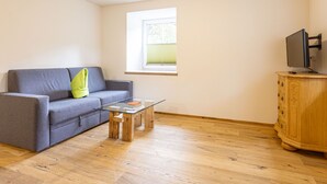 Una sala de estar con un sofá gris, una mesa de centro de vidrio, un mueble de madera y un televisor sobre un pedestal.