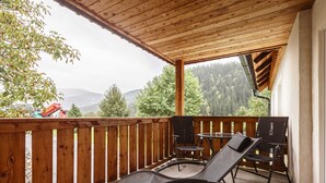 Un balcón de madera con dos sillones negros y una mesita, con vista a una zona montañosa boscosa.