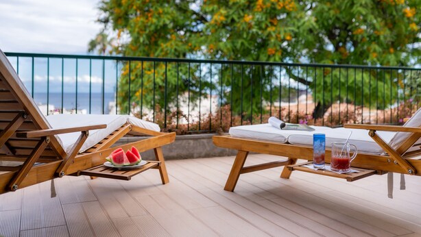 Een houten terras met twee loungesteunen, een dienblad met watermeloenpartjes en een glazen karaf met rode drank.
