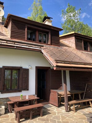 Une maison rustique en bois, dotée d’un patio en pierre, de bancs en bois et d’une table sur laquelle se trouvent une bouteille et un verre.
