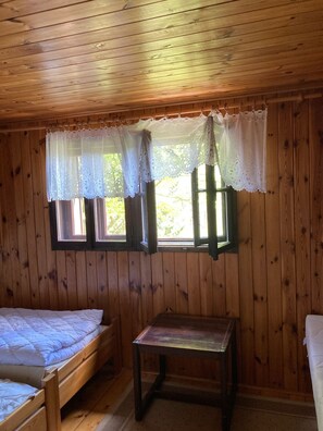 Une chambre dans un chalet en bois, avec deux lits, une petite table, des rideaux en dentelle et des fenêtres donnant sur un paysage verdoyant.