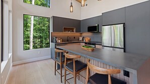 Une cuisine moderne dotée d’un îlot central, de tabourets de bar et de grandes fenêtres offrant une vue sur la forêt.