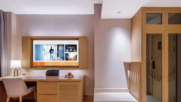 Quarto de hotel com escrivaninha de madeira, uma cadeira, televisão fixada na parede e guarda-roupa com portas de vidro.