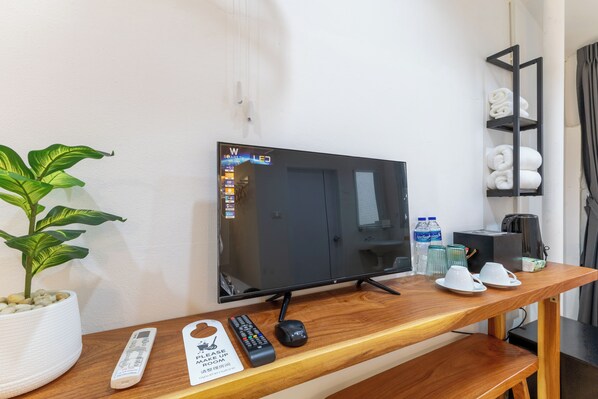 Une chambre d’hôtel avec un bureau en bois, une télévision, une télécommande, un panneau, une plante et une étagère avec des serviettes.