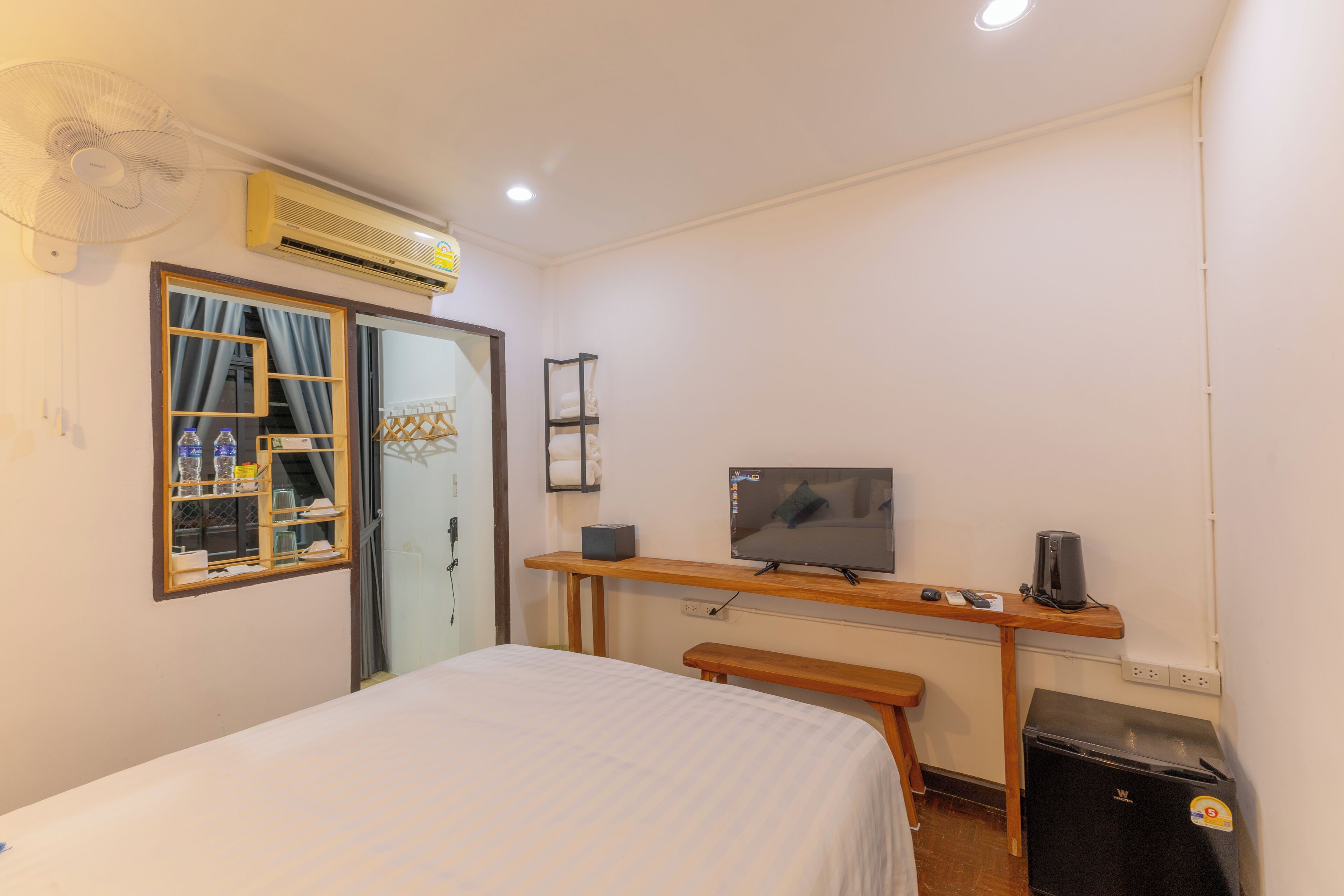 Un dormitorio con cama, escritorio con computadora, televisor, ventilador y aire acondicionado.