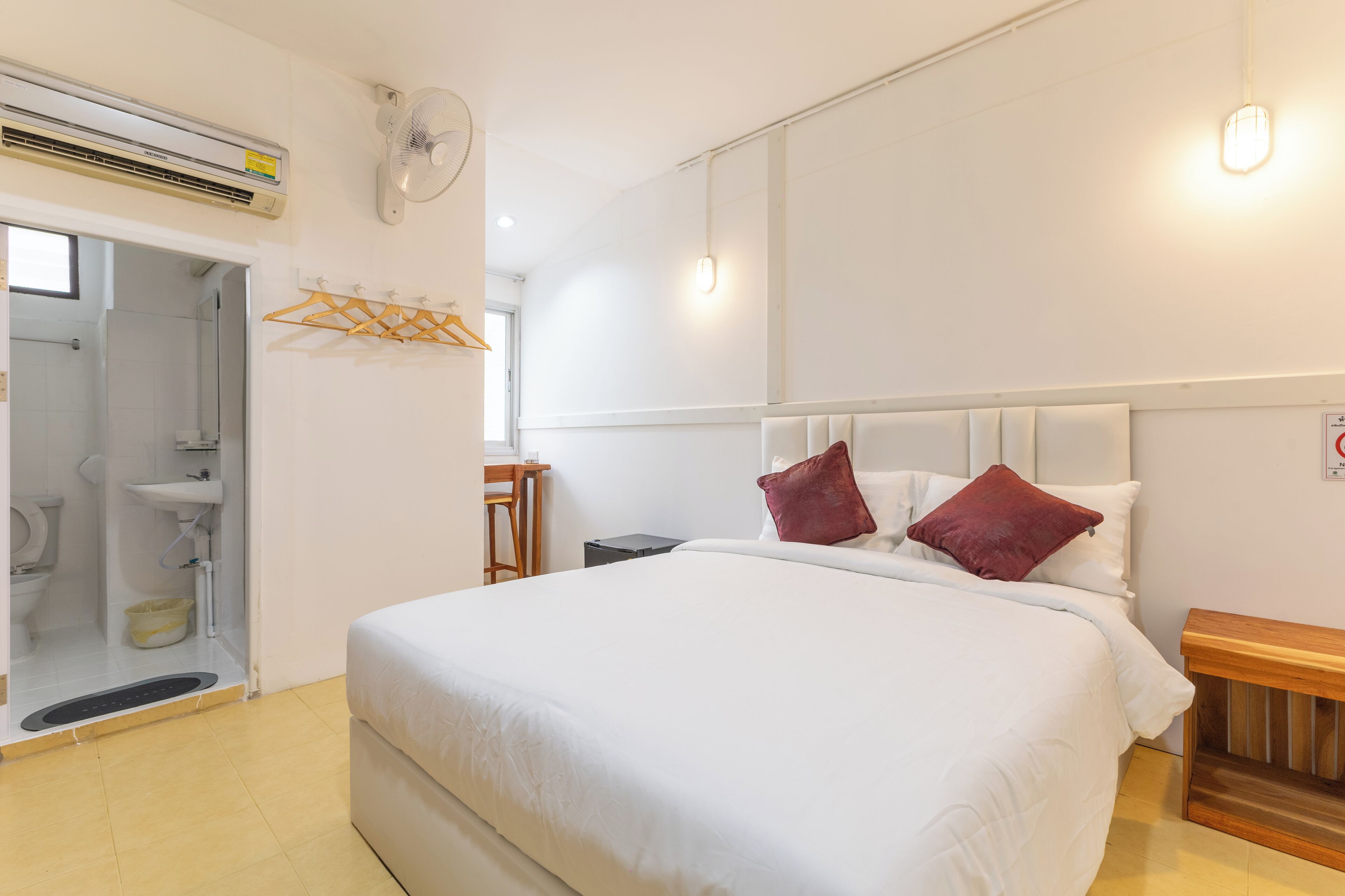 A hotel room with a bed, a wooden bedside table, a wall-mounted heater, a ceiling fan, a window, and a bathroom with a toilet and sink.