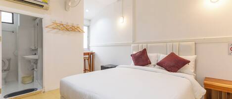 A hotel room with a bed, a wooden bedside table, a wall-mounted heater, a ceiling fan, a window, and a bathroom with a toilet and sink.