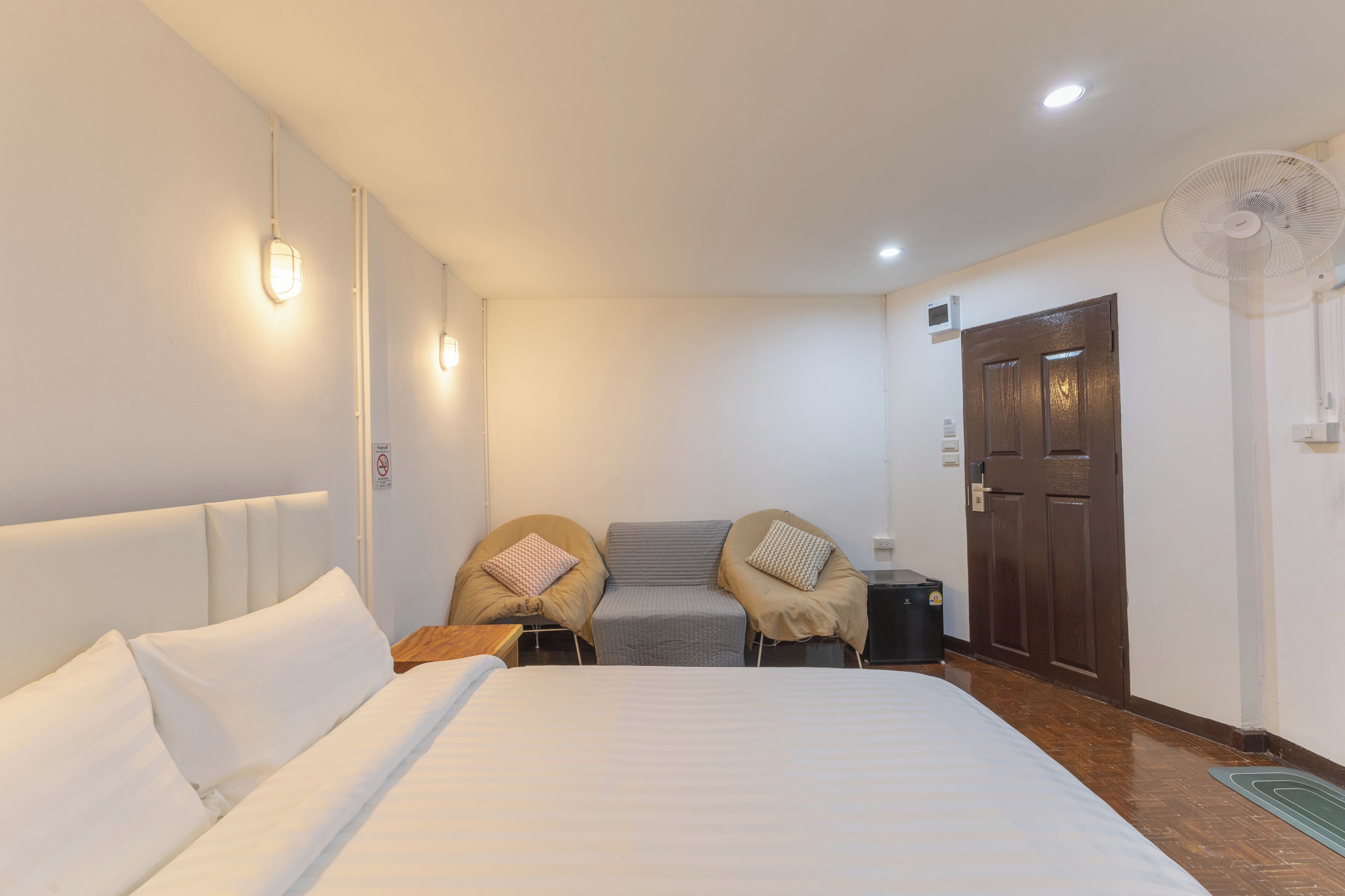 Quarto de hotel com uma cama grande, duas cadeiras, uma mesa pequena, um ventilador e uma porta.
