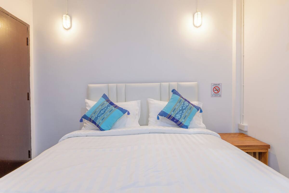 A neatly made bed with white bedding and two decorative pillows, a wooden nightstand, and a wall-mounted light fixture.