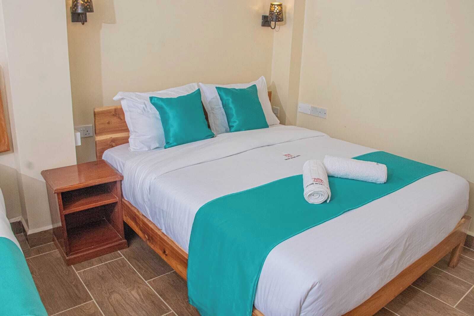 Un lit bien fait, recouvert d’une couvre-lit turquoise et blanche, avec une table de chevet en bois et une serviette blanche roulée.