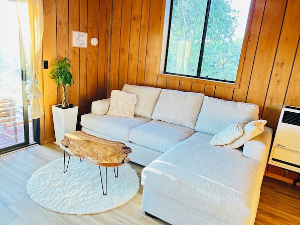 Un salon moderne avec un canapé gris clair, une table basse en bois, un tapis blanc, une plante en pot et une grande fenêtre donnant sur des arbres.