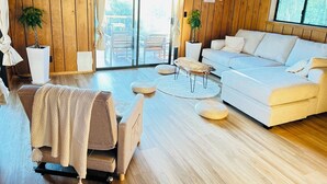 A modern living room with a sofa, a coffee table, and a TV mounted on the wall. There are two potted plants and a sliding glass door leading to an outdoor area.