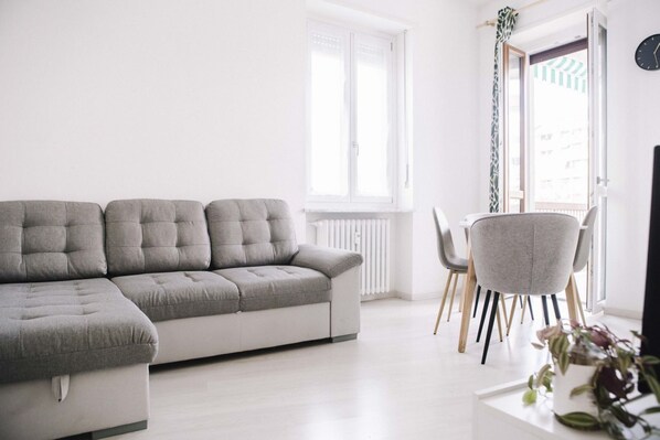 Ein modernes Wohnzimmer mit einer grauen Eckcouch, einem Esstisch mit Stühlen und einem Fenster mit Vorhängen.
