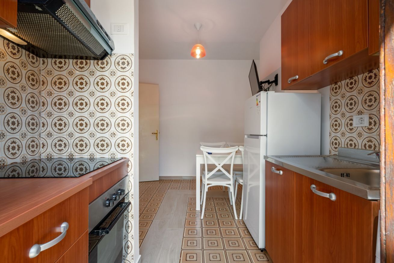 A compact kitchen with patterned tiles, a white chair, and wooden cabinets.
