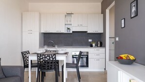 A modern kitchen with white cabinets, a dining table with chairs, and a countertop with a bowl of fruit. - GuestHost - Charming 40 sqm apartment with seaview, able to accommodate 4 people, located inside the renowned Borgo 84 residence, equipped with swimming pool and cycle path. The property is located just 3 km from the center of Marzamemi, one of the most  (Pachino)