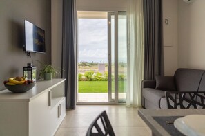 Un salon moderne doté d’un téléviseur à écran plat, d’une table à manger avec des chaises, d’un canapé et d’une vue sur un espace extérieur verdoyant, accessible par une porte-fenêtre coulissante.