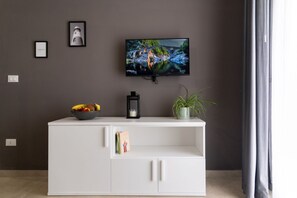 Un salon moderne avec un téléviseur à écran plat fixé au mur, un meuble de rangement blanc sur lequel sont posés un saladier de fruits et un livre, ainsi qu’une plante en pot.