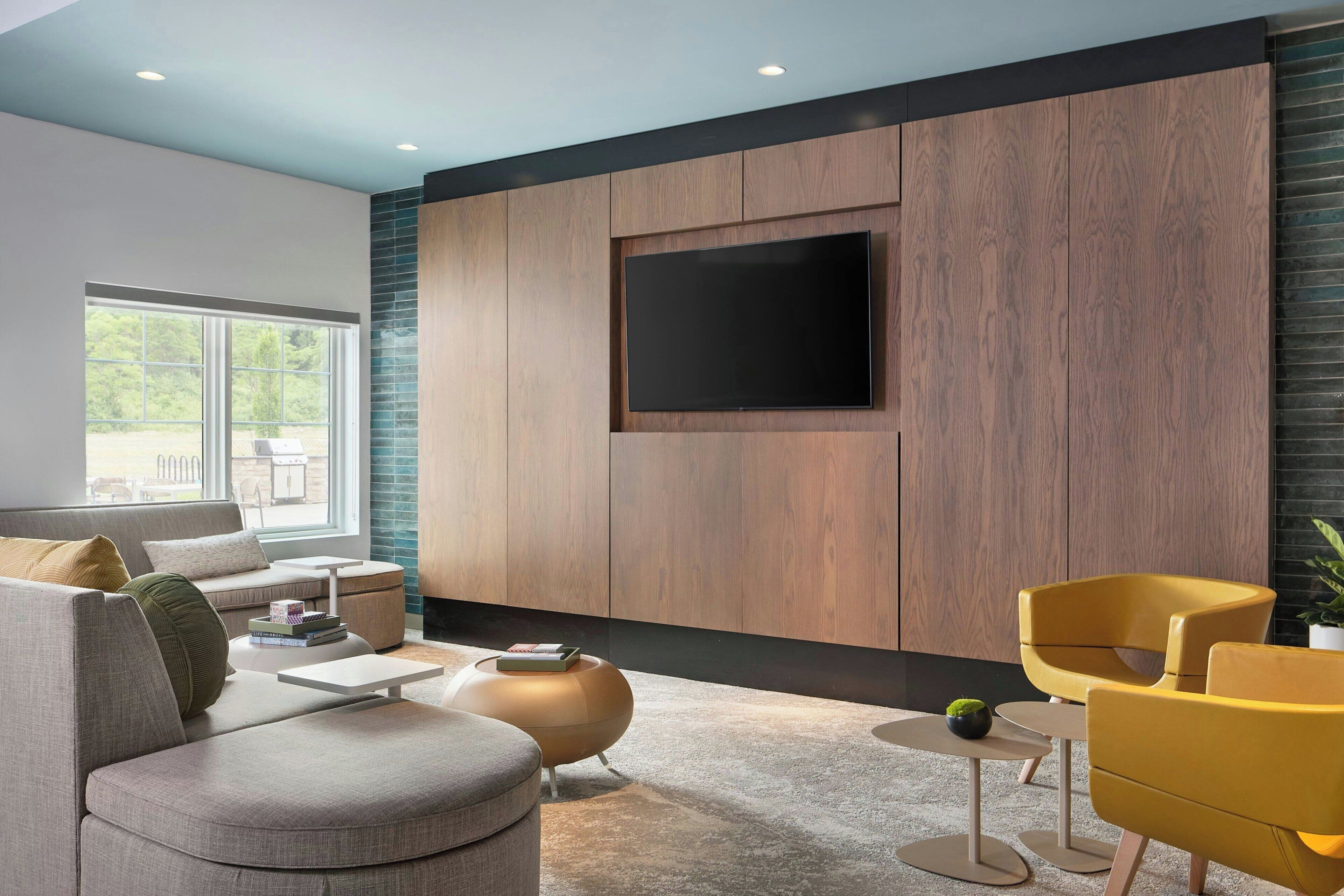 A modern living room with a large flat-screen TV mounted on a wooden wall, a gray sofa, yellow armchairs, and a small round coffee table.
