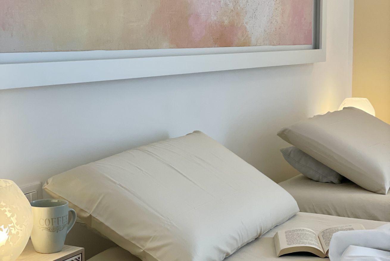 En snyggt bäddad säng med beige sängkläder och kuddar, ett sängbord med en lampa och en kaffemugg, och en inramad abstrakt målning ovanför sängen.
