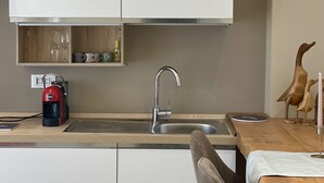 A compact kitchen with white cabinets, a stainless steel sink, a coffee maker, and wooden wall shelves.