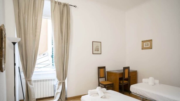 A hotel room with two single beds, a desk with a chair, a radiator, and a window with curtains. - Room Nocciola - Affitti Brevi Italia (Genova)