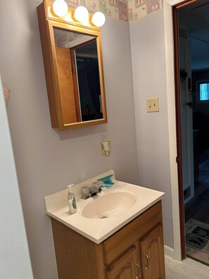 Ein Badezimmer mit einem Holzkosmetikschrank, einem Waschbecken und einem Spiegel mit Beleuchtung. An der Wand befindet sich ein Halter für eine Tasse, und eine Tür führt in einen weiteren Raum.