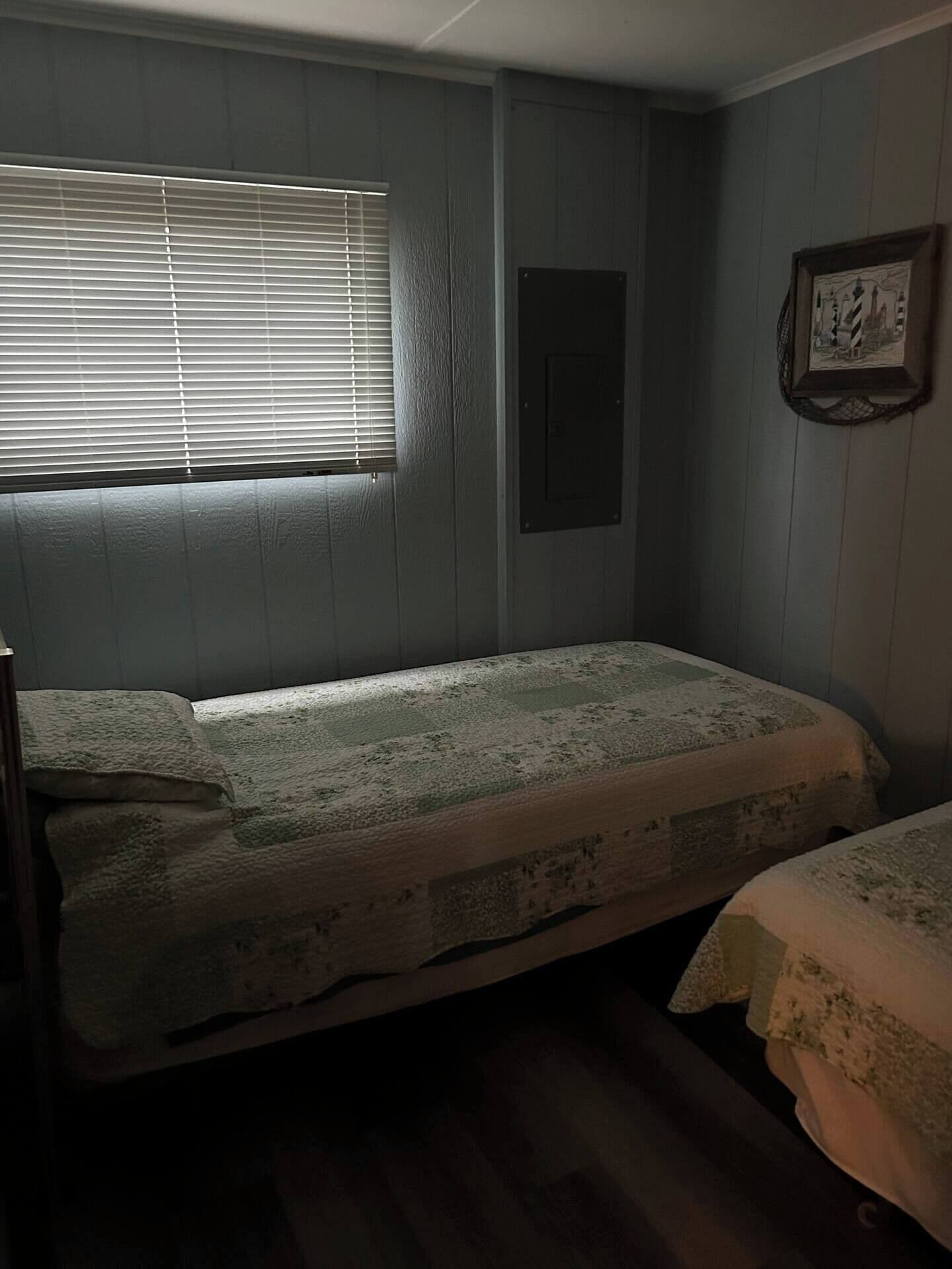 A room with two single beds, a window with blinds, a framed picture on the wall, and a small electrical panel.