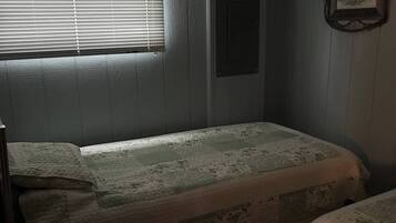 A room with two single beds, a window with blinds, a framed picture on the wall, and a small electrical panel.